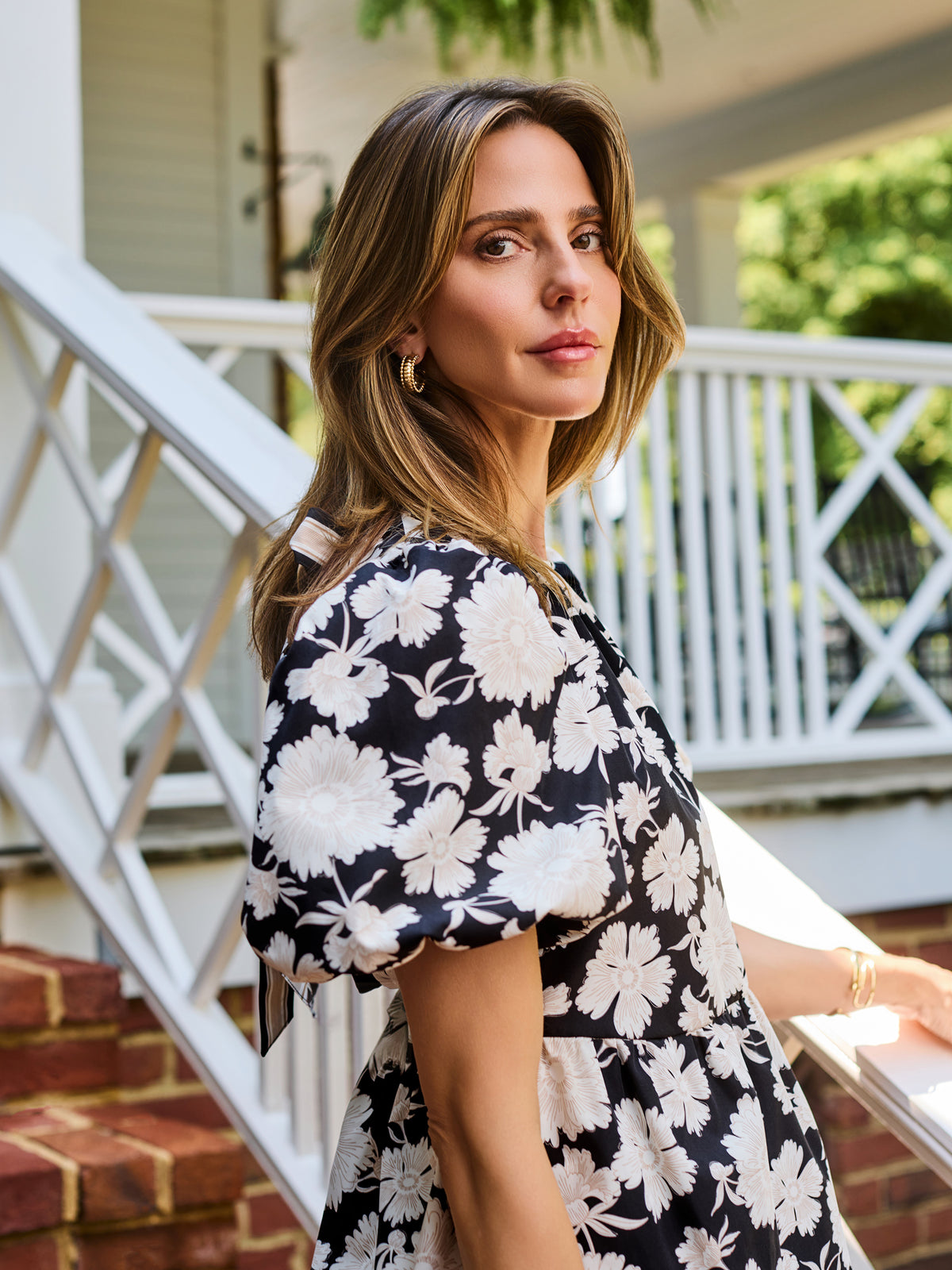 A woman wearing a black and white floral puff-sleeve  top, standing on outdoor brick steps in front of a white porch railing.