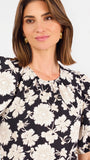 A woman modeling a black puff-sleeve blouse with a large white floral pattern, paired with blue jeans, against a plain white studio background.