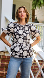 A woman stands on a porch wearing a black puff-sleeve blouse with a large white floral pattern and blue jeans.