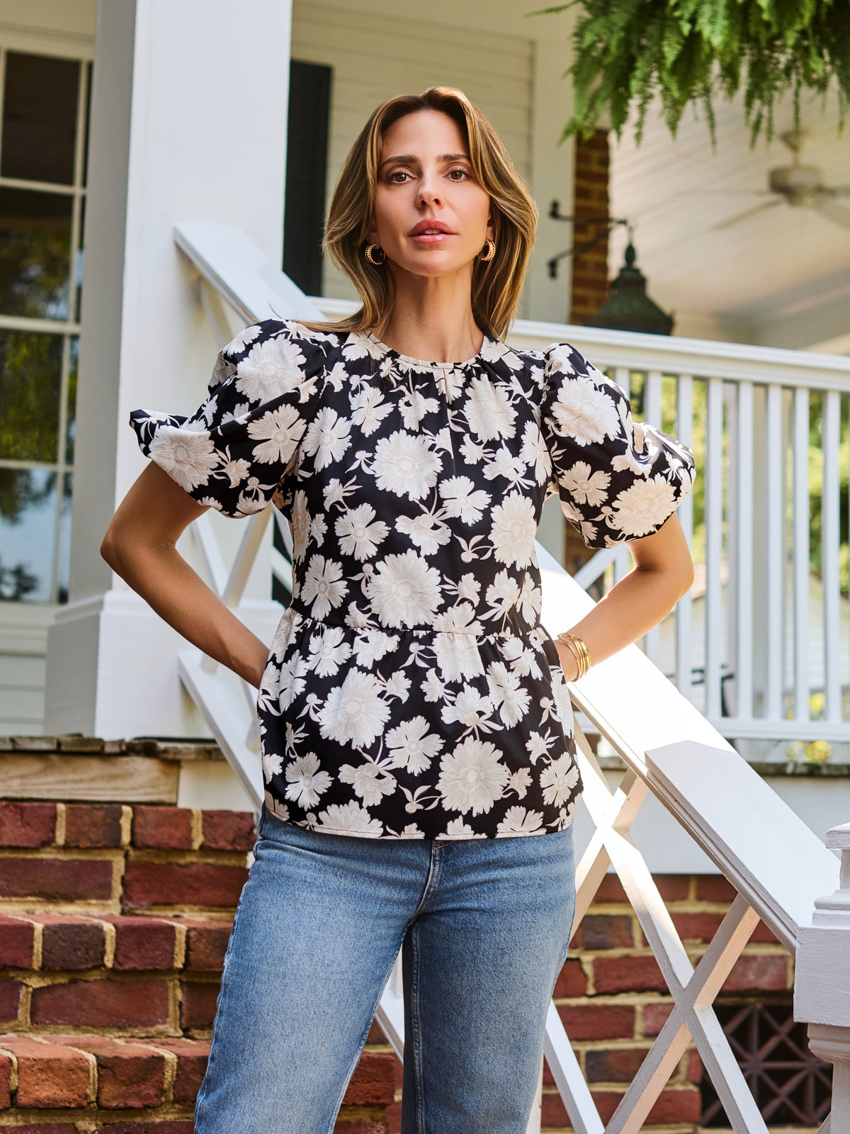 A woman stands on a porch wearing a black puff-sleeve blouse with a large white floral pattern and blue jeans.