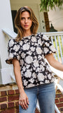 A woman wearing a black and white floral puff-sleeve  top, standing on outdoor brick steps in front of a white porch railing.