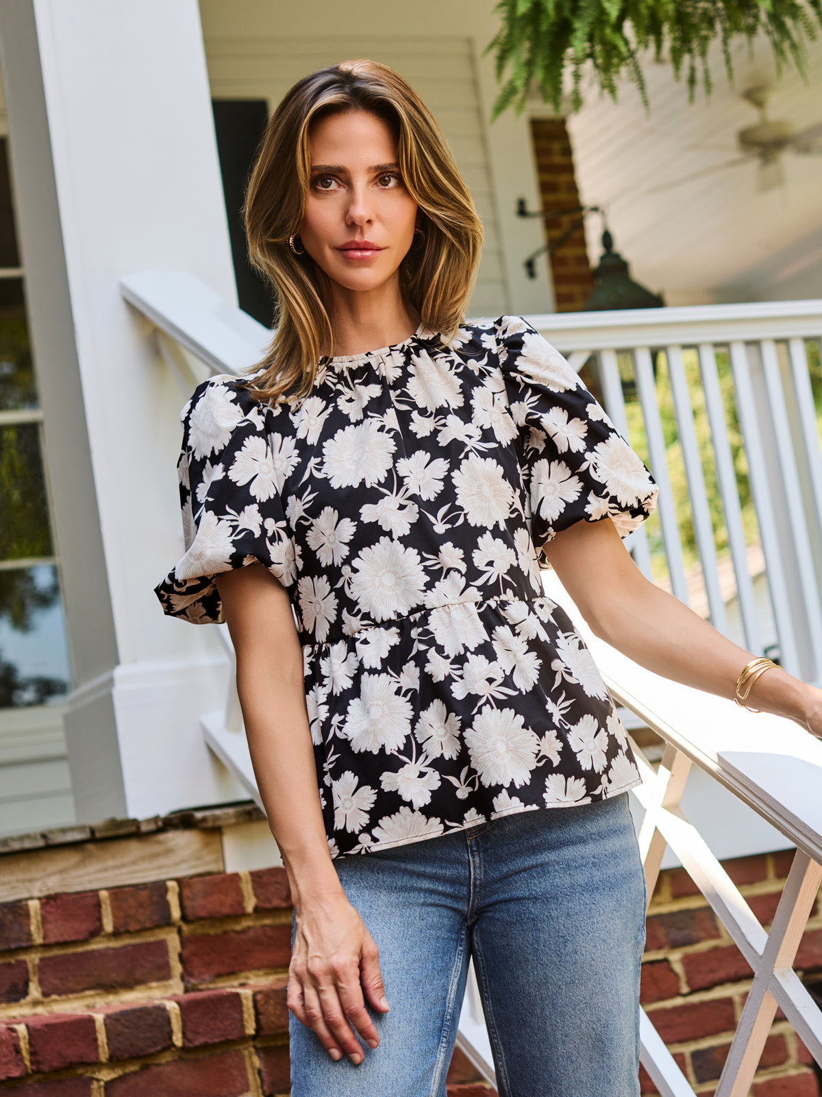 A woman wearing a black and white floral puff-sleeve  top, standing on outdoor brick steps in front of a white porch railing.