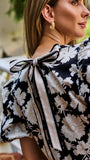 A woman wearing a black and white floral puff-sleeve  top, standing on outdoor brick steps in front of a white porch railing.