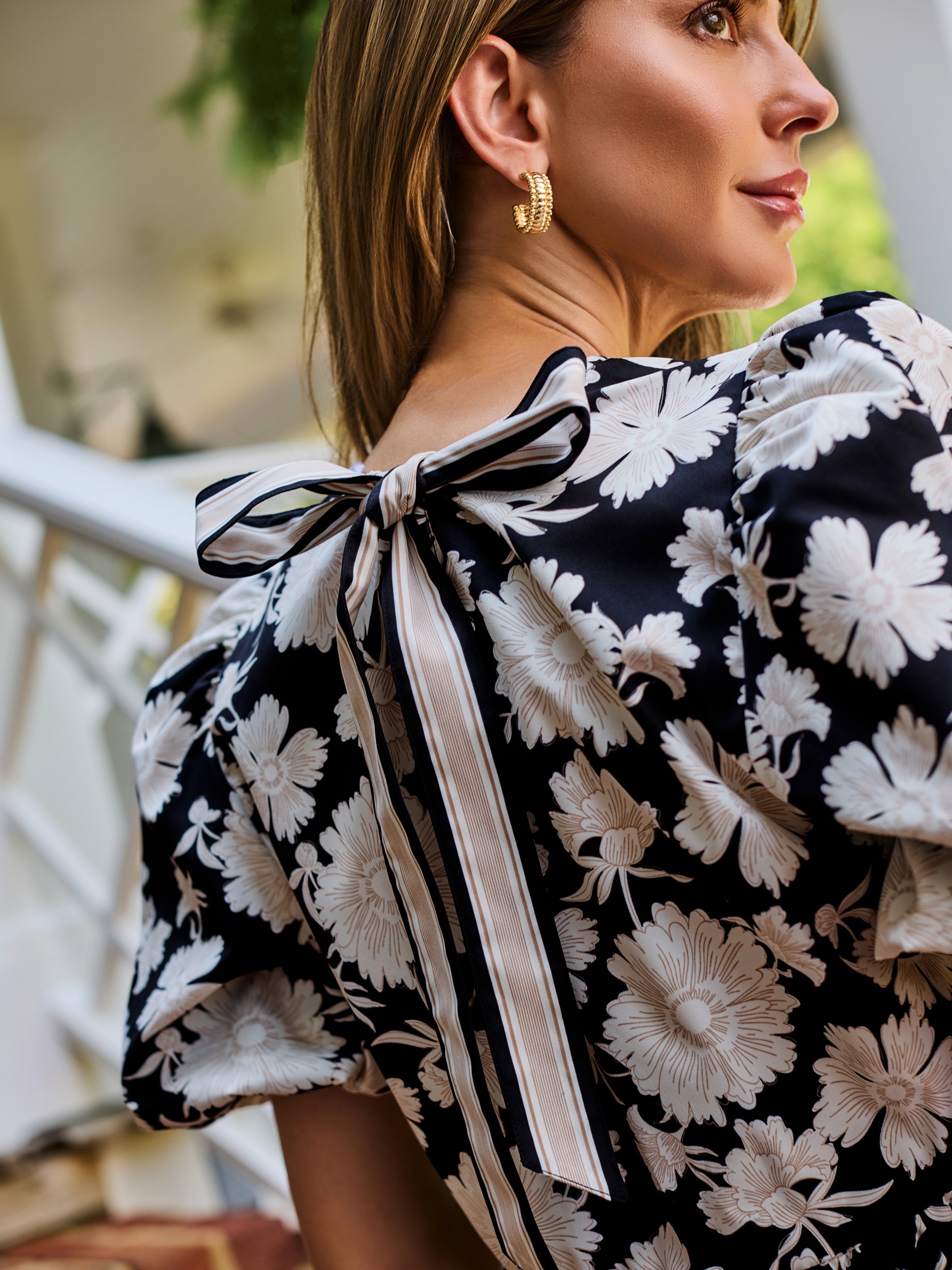 A woman wearing a black and white floral puff-sleeve  top, standing on outdoor brick steps in front of a white porch railing.