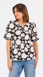 A woman modeling a black puff-sleeve blouse with a large white floral pattern, paired with blue jeans, against a plain white studio background.