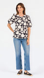 A woman modeling a black puff-sleeve blouse with a large white floral pattern, paired with blue jeans, against a plain white studio background.
