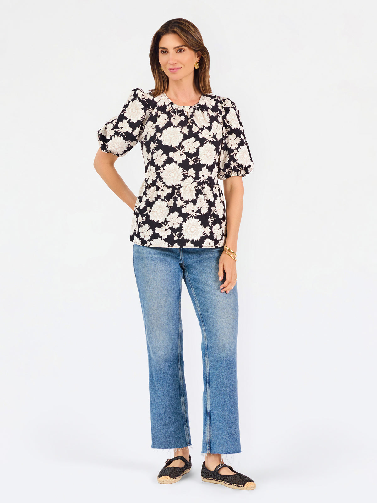 A woman modeling a black puff-sleeve blouse with a large white floral pattern, paired with blue jeans, against a plain white studio background.