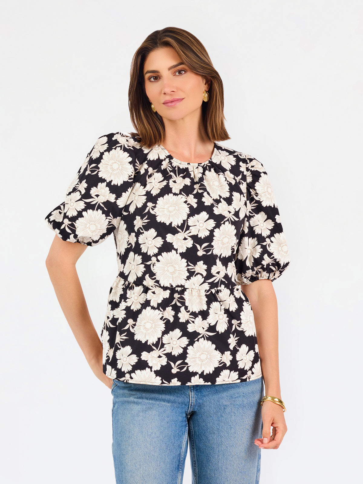 A woman modeling a black puff-sleeve blouse with a large white floral pattern, paired with blue jeans, against a plain white studio background.