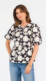 A woman modeling a black puff-sleeve blouse with a large white floral pattern, paired with blue jeans, against a plain white studio background.