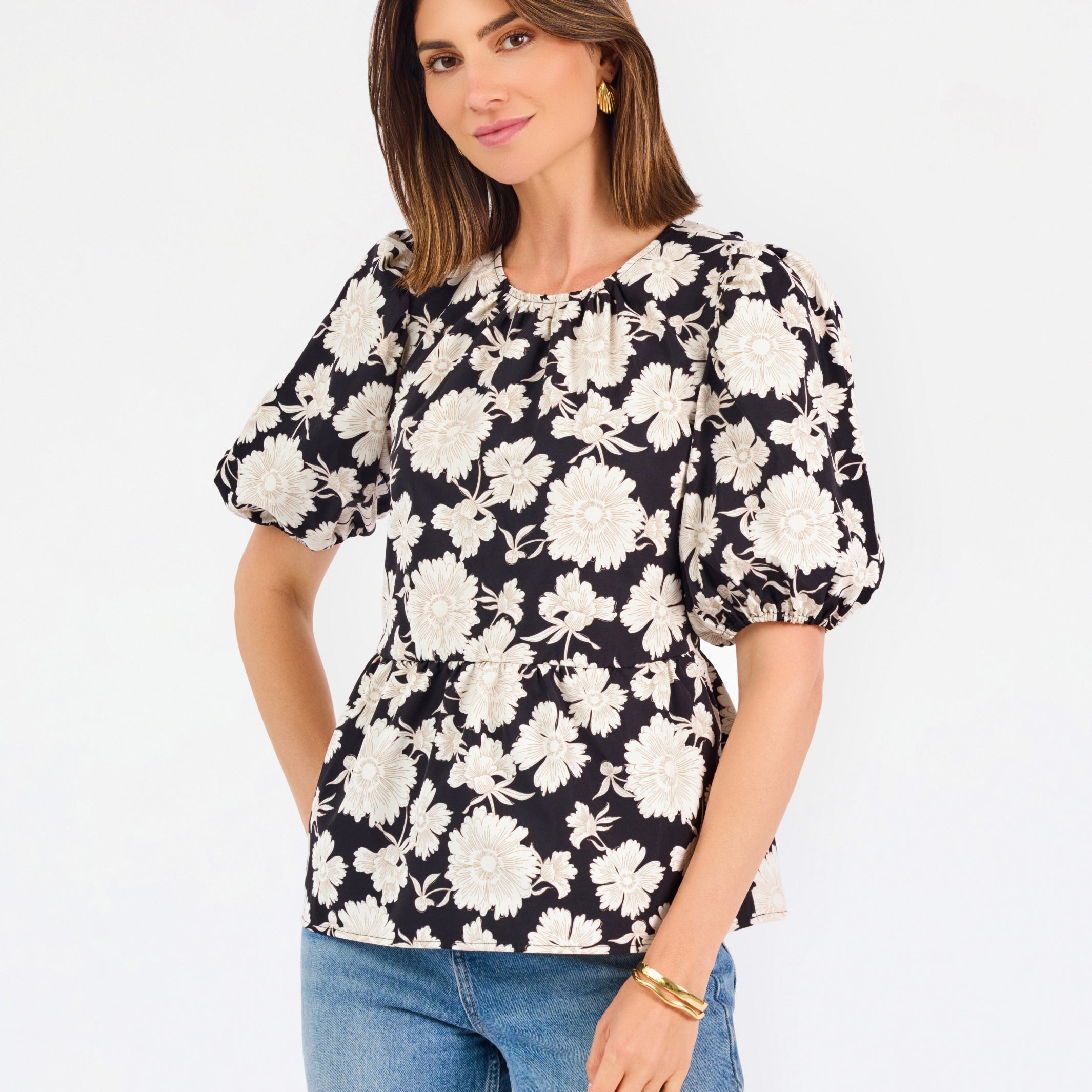 A woman modeling a black puff-sleeve blouse with a large white floral pattern, paired with blue jeans, against a plain white studio background.