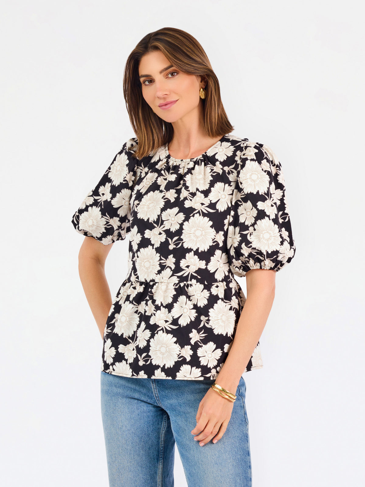 A woman modeling a black puff-sleeve blouse with a large white floral pattern, paired with blue jeans, against a plain white studio background.