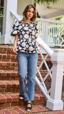 A woman wearing a black and white floral puff-sleeve  top, standing on outdoor brick steps in front of a white porch railing.