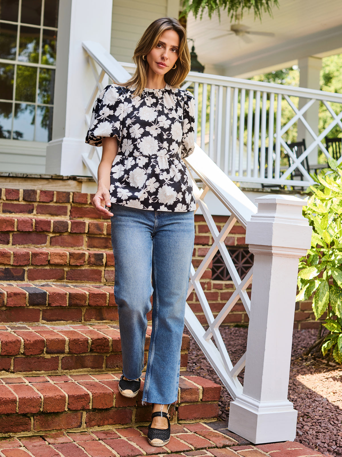 A woman wearing a black and white floral puff-sleeve  top, standing on outdoor brick steps in front of a white porch railing.
