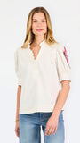 A woman wearing a cream-colored blouse with a ruffled collar, short puff sleeves, and pink floral embroidery on the upper left sleeve, shown against a plain white background.