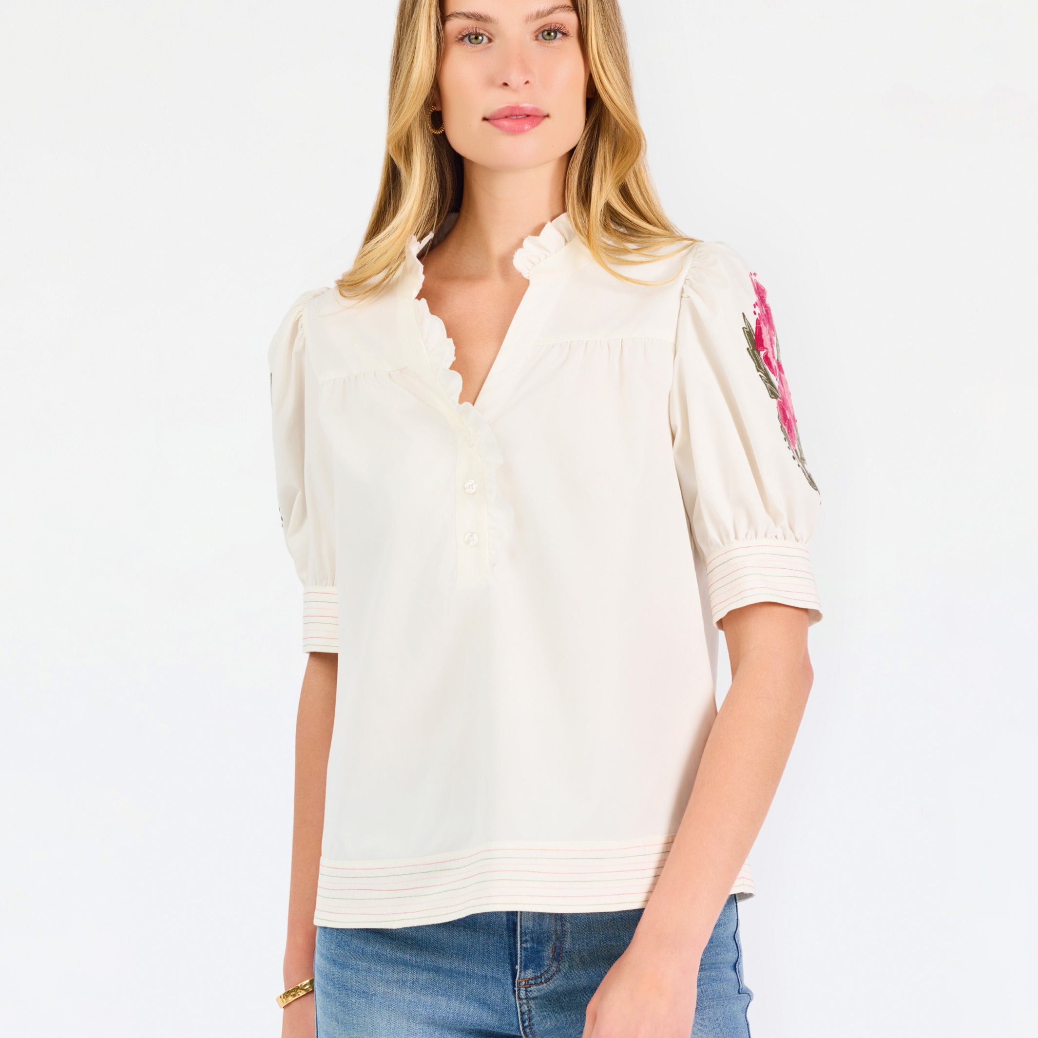 A woman wearing a cream-colored blouse with a ruffled collar, short puff sleeves, and pink floral embroidery on the upper left sleeve, shown against a plain white background.