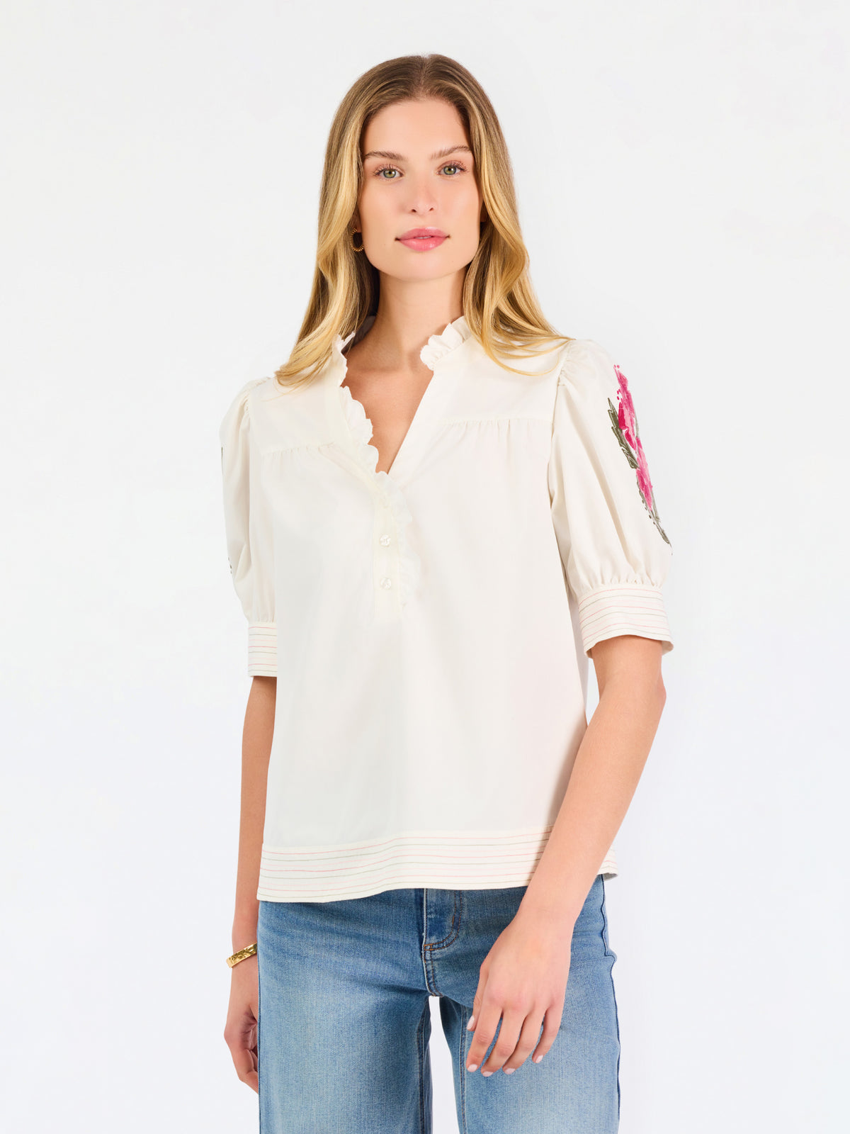 A woman wearing a cream-colored blouse with a ruffled collar, short puff sleeves, and pink floral embroidery on the upper left sleeve, shown against a plain white background.