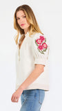 A woman wearing a cream-colored blouse with a ruffled collar, short puff sleeves, and pink floral embroidery on the upper left sleeve, shown against a plain white background.