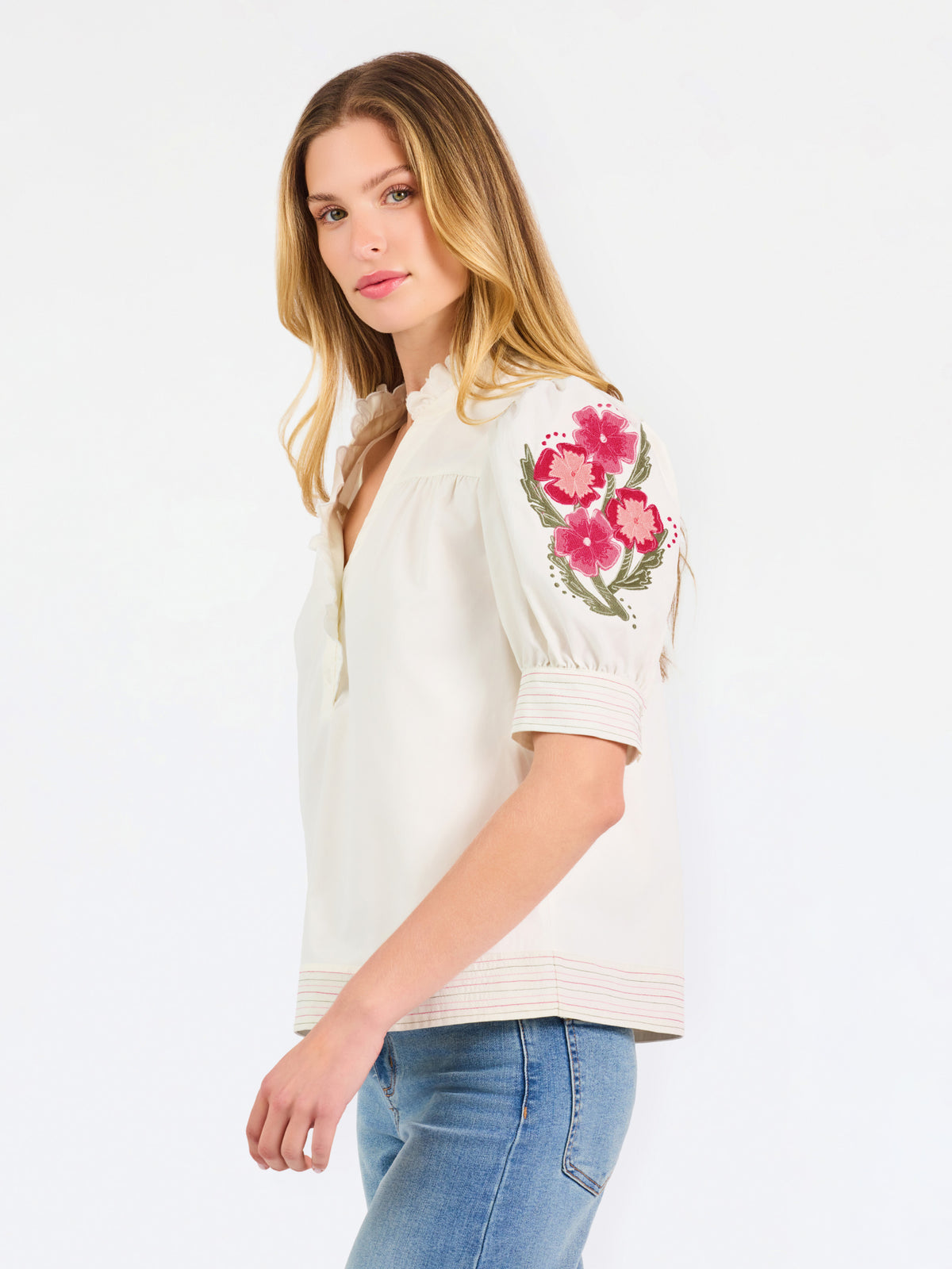 A woman wearing a cream-colored blouse with a ruffled collar, short puff sleeves, and pink floral embroidery on the upper left sleeve, shown against a plain white background.