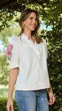 A woman wearing a cream-colored short-sleeve blouse with pink floral embroidery on the sleeve and a ruffled neckline, paired with blue jeans, standing outdoors in front of lush green foliage.