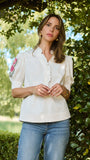 A woman wearing a white short-sleeve blouse with floral embroidery on the sleeve and blue jeans, standing outdoors in front of a leafy green hedge.