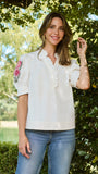 A woman wearing a white short-sleeve blouse with floral embroidery on the sleeve and ruffled trim, paired with blue jeans, standing outdoors in front of green foliage.