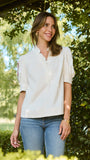 A woman wearing a short-sleeve cream blouse with puff sleeves and a ruffled collar, paired with blue jeans, standing outdoors in front of a leafy green hedge.