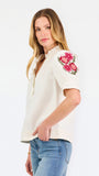 A woman wearing a cream-colored blouse with a ruffled collar, short puff sleeves, and pink floral embroidery on the upper left sleeve, shown against a plain white background.
