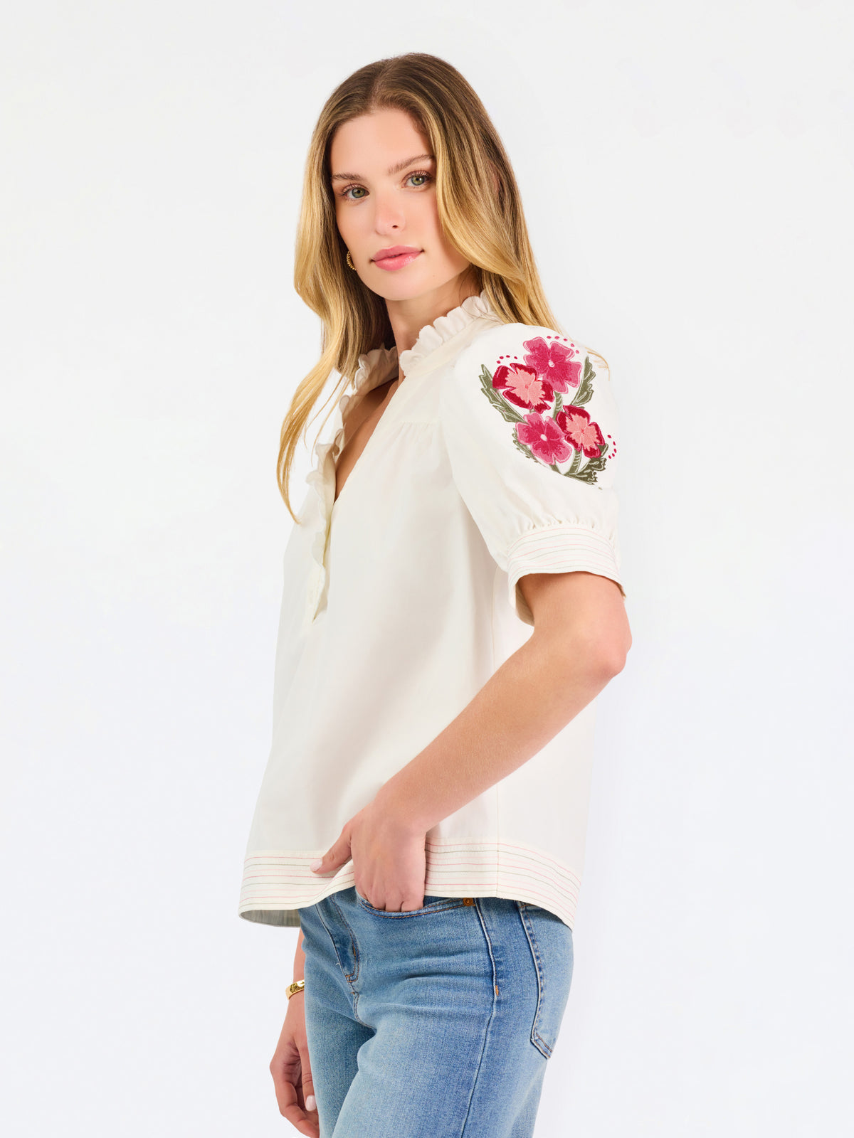 A woman wearing a cream-colored blouse with a ruffled collar, short puff sleeves, and pink floral embroidery on the upper left sleeve, shown against a plain white background.