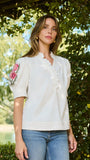 A woman wearing a white short-sleeve blouse with floral embroidery on the sleeve and ruffled trim, paired with blue jeans, standing outdoors under a leafy garden arch.