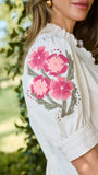 A woman wearing a white blouse with pink floral embroidery on the sleeve, photographed outdoors against a blurred green foliage background.