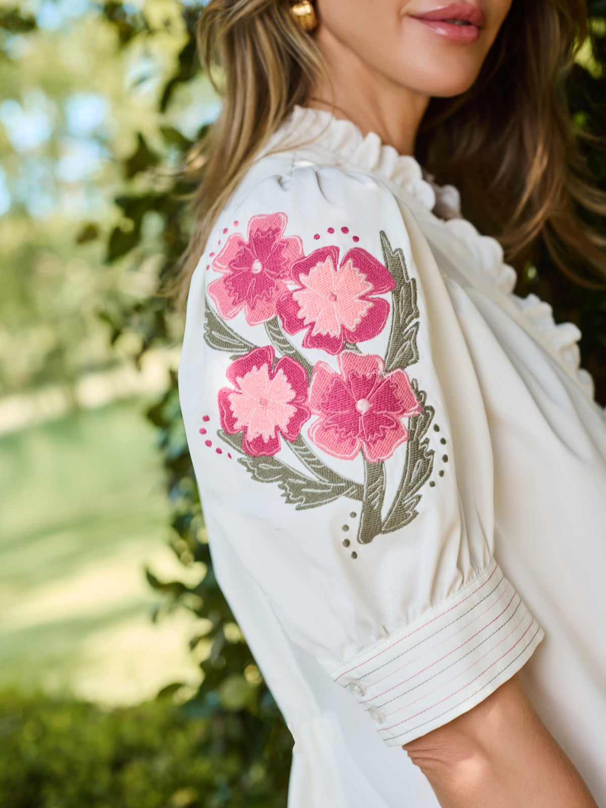 A woman wearing a white blouse with pink floral embroidery on the sleeve, photographed outdoors against a blurred green foliage background.