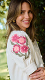 A woman wearing a white blouse with pink floral embroidery on the sleeve, photographed outdoors against a blurred green foliage background.