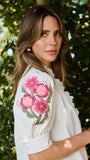A woman wearing a white blouse with pink floral embroidery on the sleeve, photographed outdoors against a blurred green foliage background.