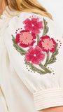 A woman wearing a cream-colored blouse with a ruffled collar, short puff sleeves, and pink floral embroidery on the upper left sleeve, shown against a plain white background.
