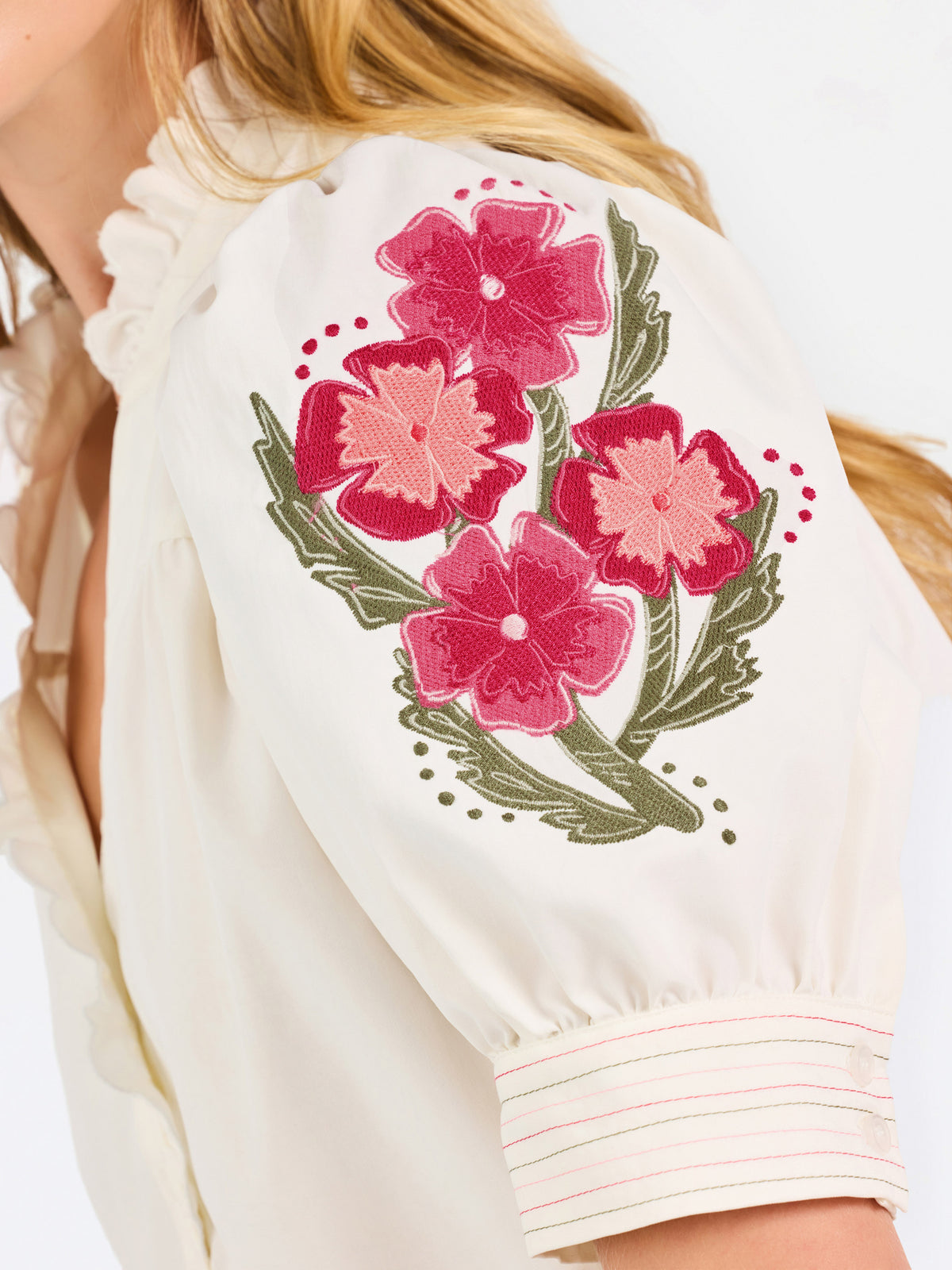 A woman wearing a cream-colored blouse with a ruffled collar, short puff sleeves, and pink floral embroidery on the upper left sleeve, shown against a plain white background.