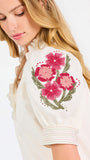 A woman wearing a cream-colored blouse with a ruffled collar, short puff sleeves, and pink floral embroidery on the upper left sleeve, shown against a plain white background.