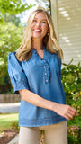 A smiling blonde woman wearing a blue short-sleeve button-front blouse with ruffle trim and beige pants, standing outdoors in front of green bushes and a house wall.