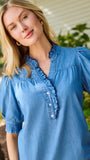 A smiling blonde woman wearing a blue short-sleeve button-front blouse with ruffle trim and beige pants, standing outdoors in front of green bushes and a house wall.