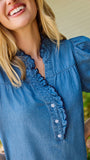 A smiling blonde woman wearing a blue short-sleeve button-front blouse with ruffle trim and beige pants, standing outdoors in front of green bushes and a house wall.