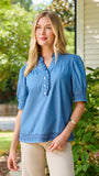 A smiling blonde woman wearing a blue short-sleeve button-front blouse with ruffle trim and beige pants, standing outdoors in front of green bushes and a house wall.