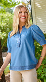 A smiling blonde woman wearing a blue short-sleeve button-front blouse with ruffle trim and beige pants, standing outdoors in front of green bushes and a house wall.