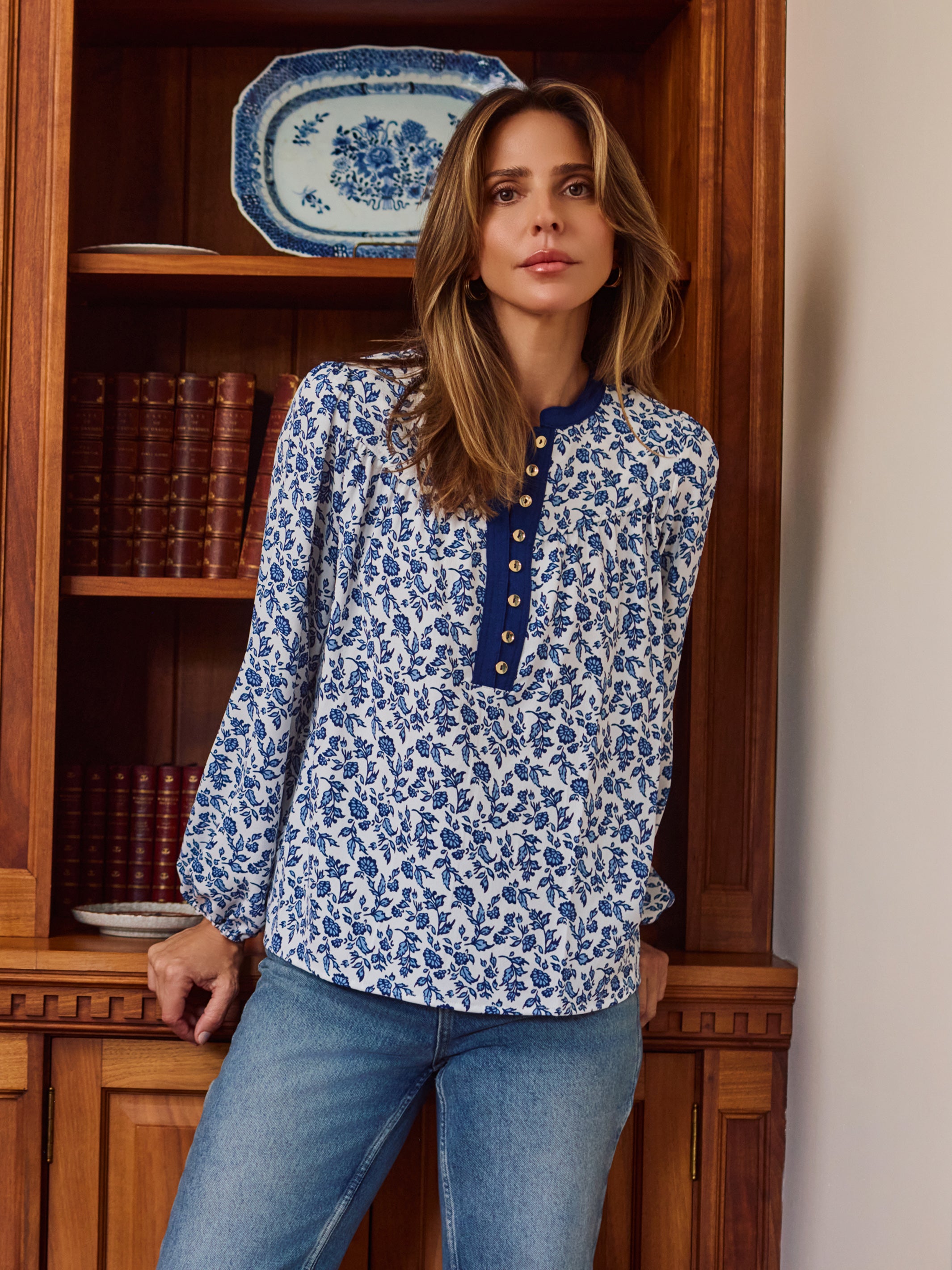 A woman wearing a blue and white floral print long-sleeve blouse with a buttoned collar, standing in front of a wooden bookshelf.