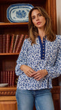 A woman wearing a blue and white floral print long-sleeve blouse with a buttoned collar, standing in front of a wooden bookshelf.