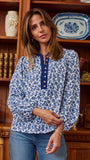 A woman wearing a blue and white floral print long-sleeve blouse with a buttoned collar, standing in front of a wooden bookshelf.
