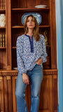 A woman wearing a blue and white floral print long-sleeve blouse with a buttoned collar, standing in front of a wooden bookshelf.