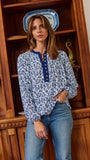 A woman wearing a blue and white floral print long-sleeve blouse with a buttoned collar, standing in front of a wooden bookshelf.