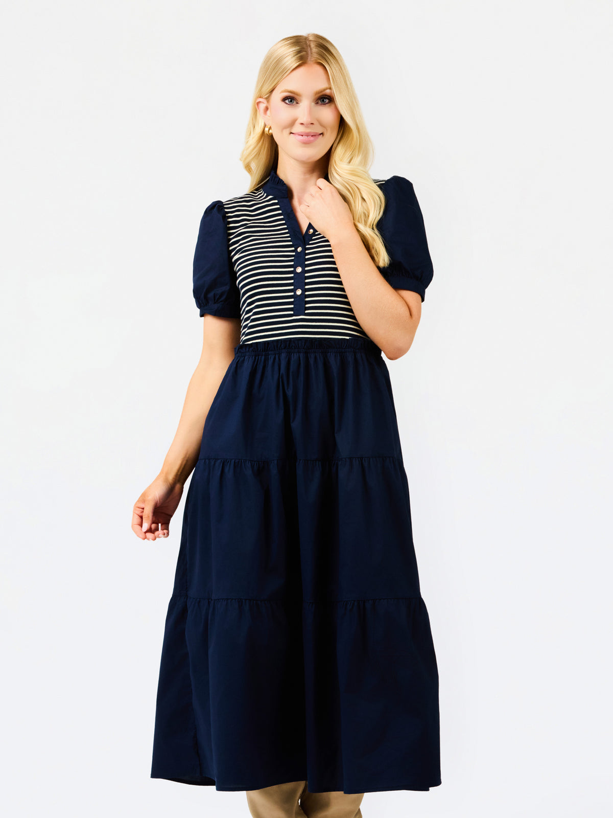 A woman modeling a navy blue tiered midi dress with a navy and white striped button-up bodice and puff sleeves, set against a plain white studio background.