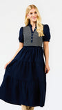 A blonde model wearing a navy blue tiered midi dress with a striped yoke and puff sleeves, posed against a solid white studio background.