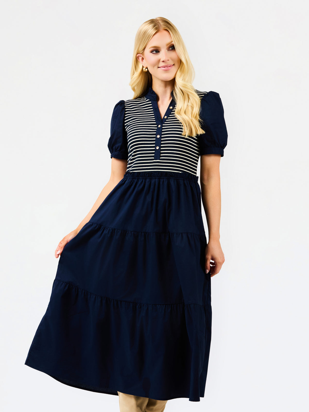 A blonde model wearing a navy blue tiered midi dress with a striped yoke and puff sleeves, posed against a solid white studio background.