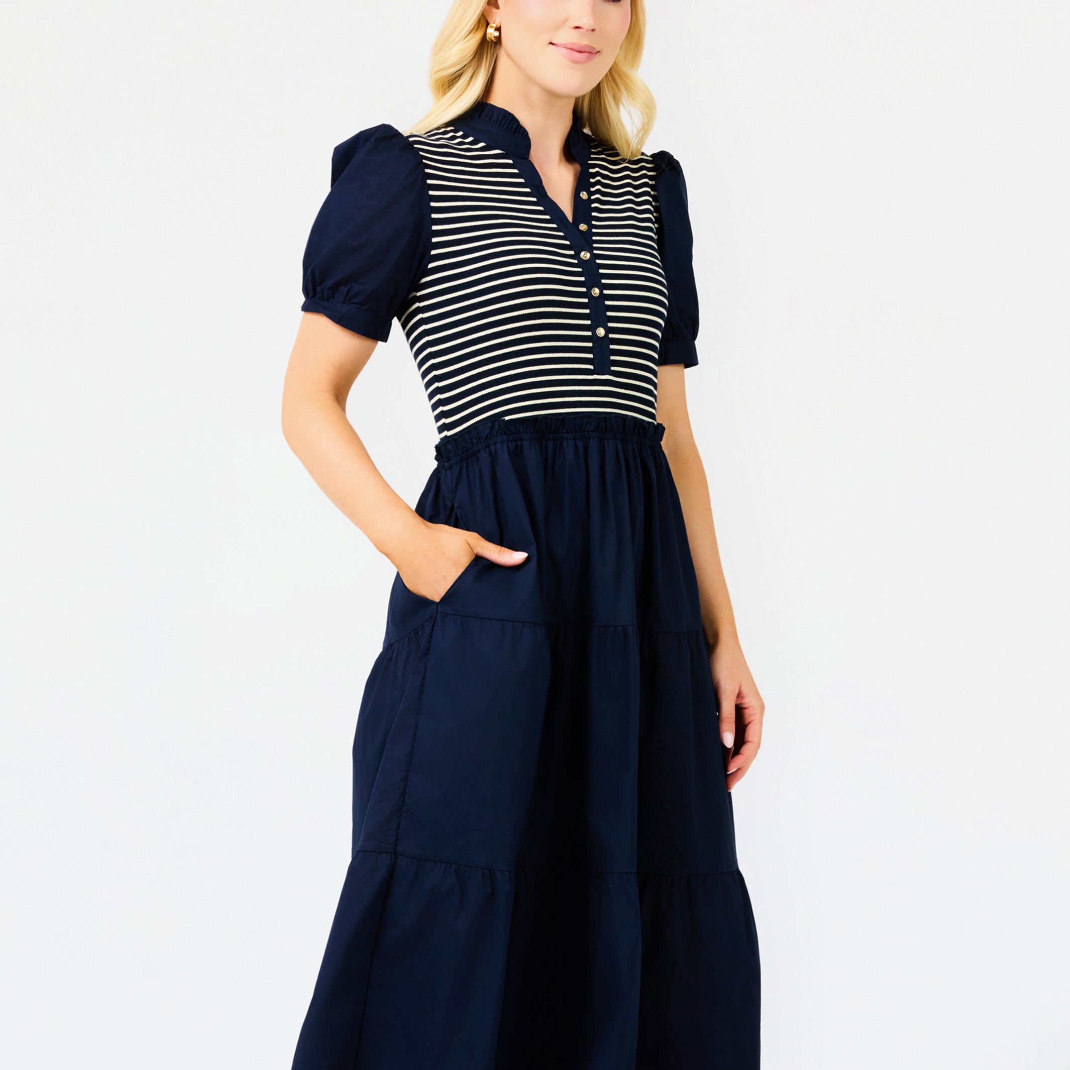 A woman modeling a navy blue midi dress with a navy and white striped bodice, short puff sleeves, and a tiered skirt, set against a plain white studio background.